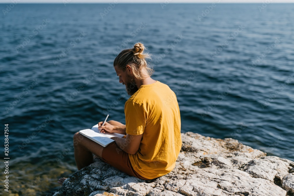 Fototapeta premium Young man with a top bun writing in a notebook on a rocky seaside by the ocean for journal background and creative retreat imagery