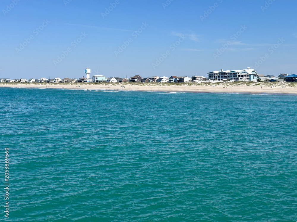 Fototapeta premium Clear turquoise waters of the Atlantic Ocean stretch toward the sandy shoreline and beachfront homes of Atlantic Beach, North Carolina on a bright summer day.