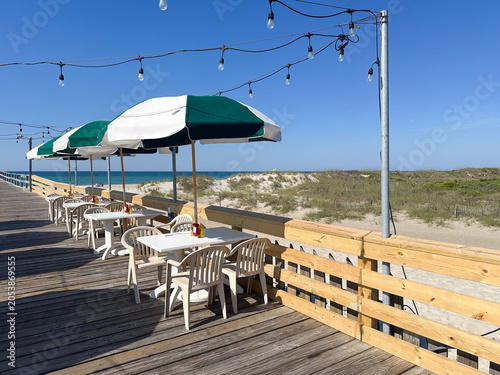 Outdoor dining tables with striped umbrellas overlook sandy dunes and the Atlantic Ocean from a beachfront boardwalk pier in Atlantic Beach, North Carolina on a sunny summer day.