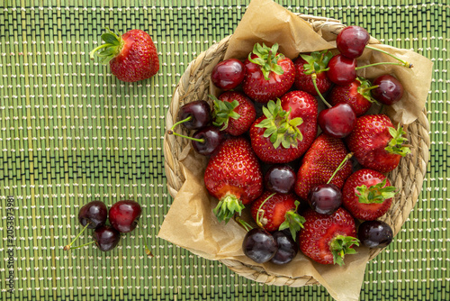 Fresh ripe red strawberries and sweet cherry in a woven basket on green placemat, top view