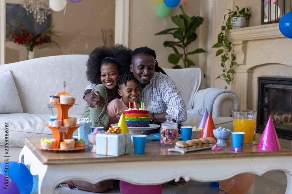 Obraz premium African American family sitting in living room, hugging child, blowing lit candles on rainbow cake