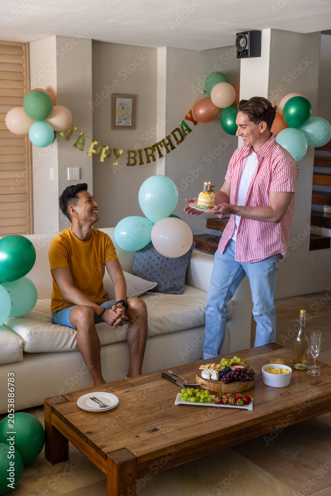 Naklejka premium Diverse men carrying cake with lit candles, wearing mustard tee and pink shirt celebrating at home