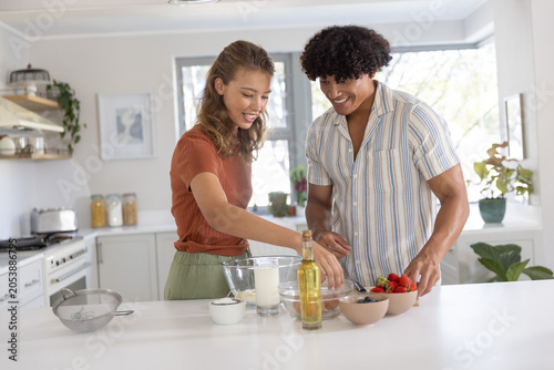 Couple mixing batter on bright white kitchen island with glass bowl, milk, berries, olive oil