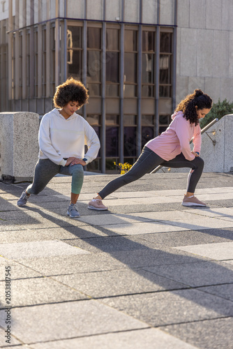 Schilderij op canvas Two adult women stretching and lunging on sunlit concrete plaza near office buil