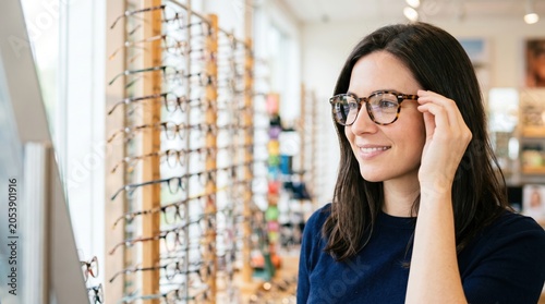 A young woman looks at herself in the mirror while trying on a pair of glasses. She stands in an optical store with many frames displayed on the wall