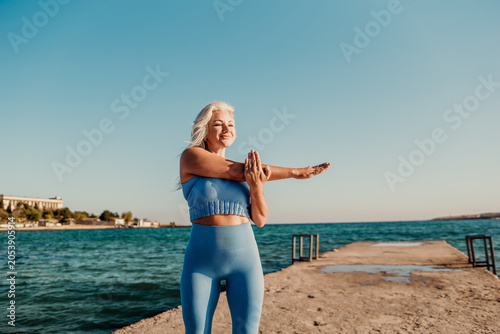 Fotografia Woman Yoga Stretching Beach - A young woman in blue sportswear stretches her arms while standing on a pier overlooking the blue sea on a sunny day