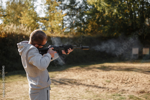 Foto Man In Gray Hoodie Firing Automatic Rifle At Outdoor Shooting Range With Ejecting Shell Case