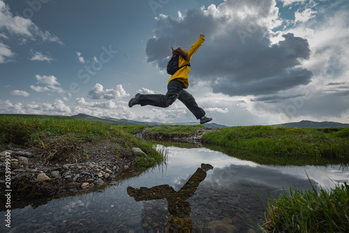 Fotografie Active woman hiker with backpack jumping high in midair over mountain stream with water reflection