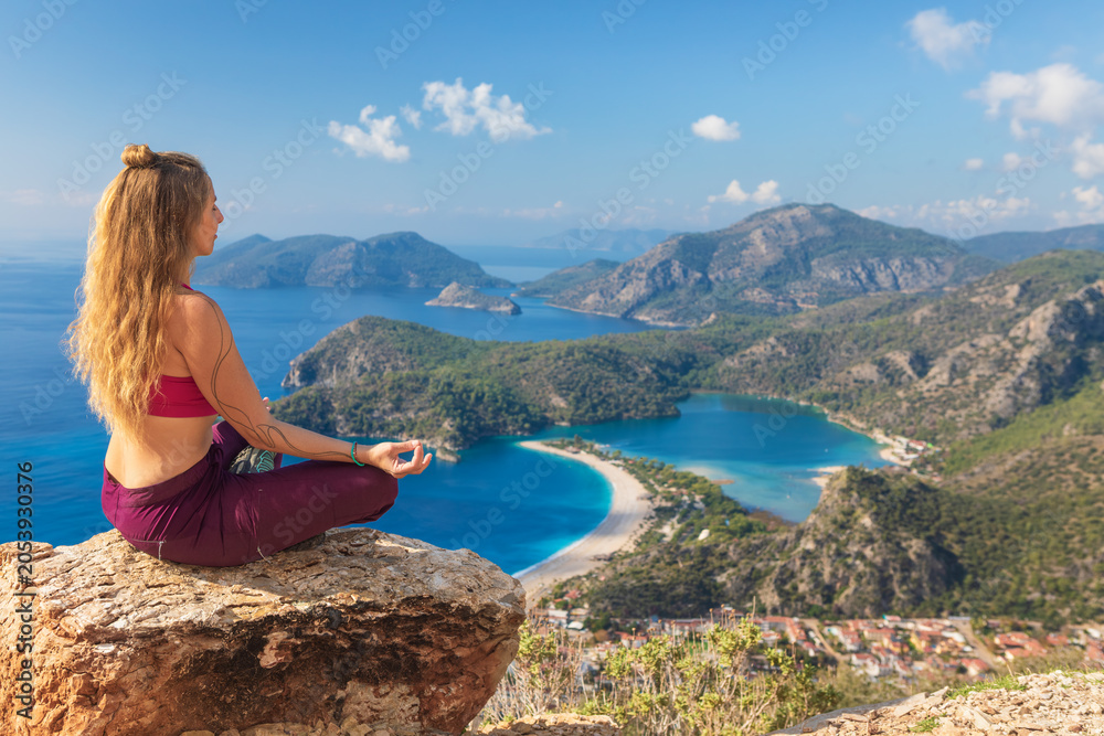 Obraz premium Young woman sit in yoga pose on rock top relaxing, looking at blue lagoon, Oludeniz beach. Summer day walk by Lycian way at family vacation in Mediterranean Sea coast, Fethye, Antalya region, Turkey