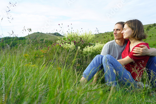 Foto Man and woman sit close in wildflower meadow, his arm around her shoulders as they gaze toward hills