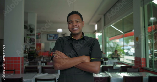 Latino restaurant employee smiling with arms crossed, cheerful and confident body language in bright indoor restaurant setting