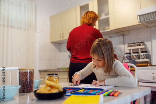 Little girl doing homework while mother making breakfast at home