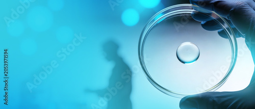 Gloved hand holding a petri dish with a liquid drop against a blurred silhouette of a scientist in blue light.