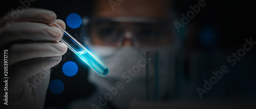 Gloved hand holding a test tube with glowing blue liquid against a dark background with a blurred laboratory worker.