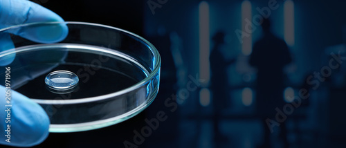 Petri dish with a gel-like sample droplet held by a hand in a blue glove against blurred silhouettes of scientists in a lab.