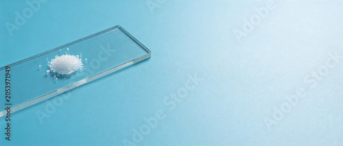 Small heap of white crystalline powder on a glass microscope slide against a light blue background.