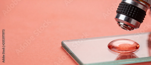 Microscope objective lens pointing at a drop of reddish liquid on a glass slide positioned on a peach colored background.