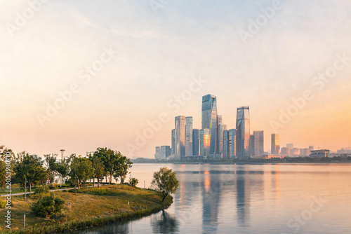 Modern skyscrapers skyline reflected on calm river at sunset with green riverside park in foreground