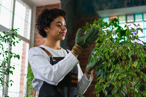 Running of own business. African woman florist wearing apron taking care of plants in botanical store. Happy small business owner working at flower shop smiling surrounded by plants Small business