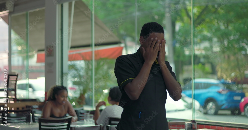 Fototapeta premium Restaurant worker covering face with both hands near window, showing signs of burnout, emotional exhaustion, and overwhelming fatigue
