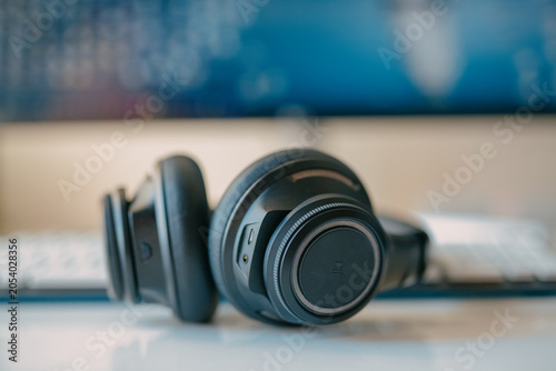 A close-up shot of a pair of modern black wireless headphones resting on a white desk, with a keyboard and a blurred blue computer screen in the soft-focus background.