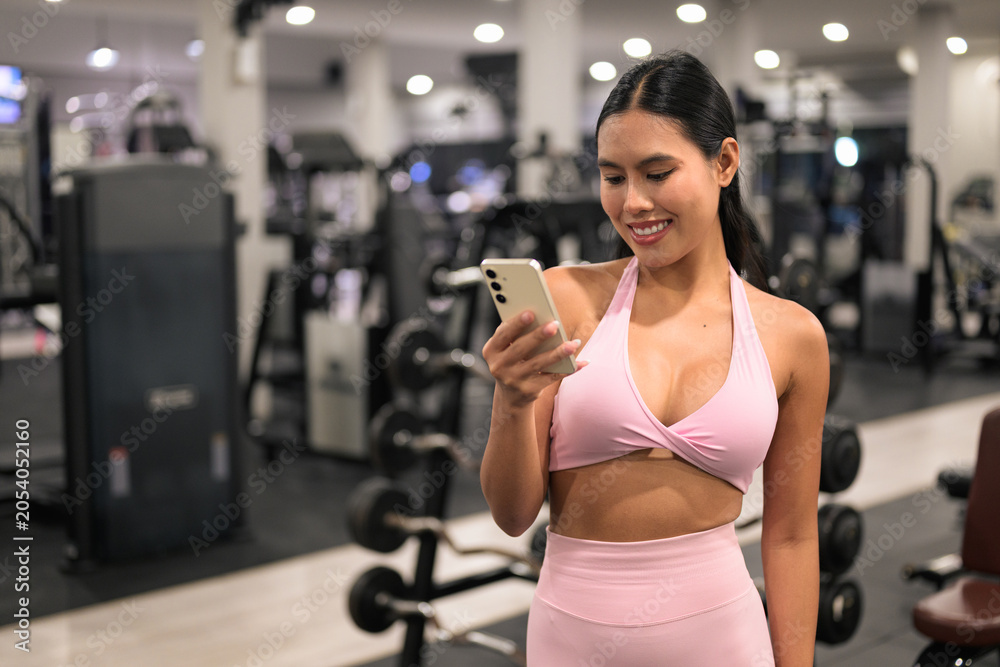 Fototapeta premium Smiling Asian woman in pink activewear using smartphone in fitness gym