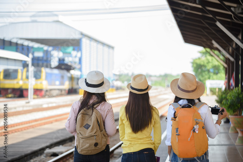 Three Asian women tourists with backpack waiting at railway station platform view train on track during summer vacation holiday travel journey lifestyle group adventure from behind