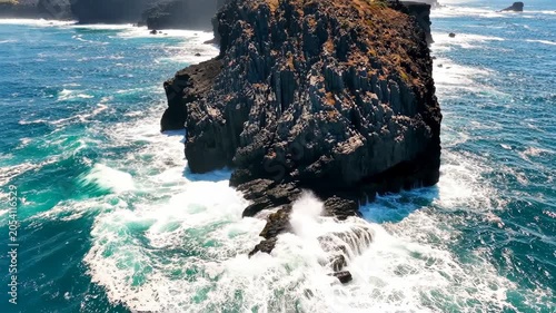 Large dark rock formation in the middle of a blue ocean with crashing white waves