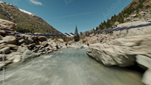 An Fpv drone flies low over a mountain river, flies under an overhead bridge. The large snow-capped peaks of the mountains are visible in the background. High quality 4k footage