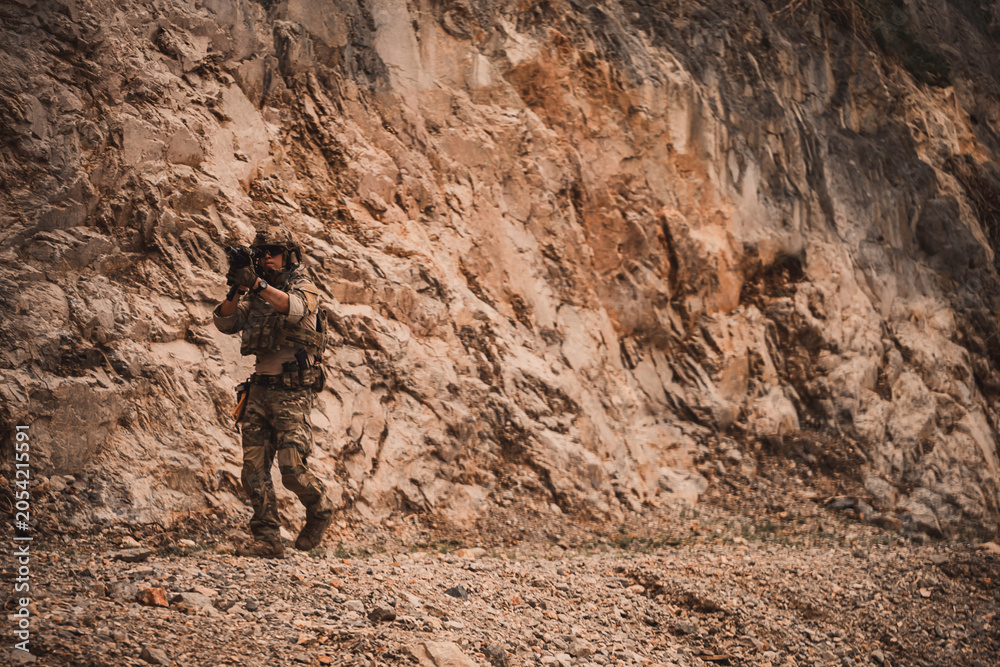 Naklejka premium Armed military operator advancing through rugged rocky terrain while aiming a rifle during a combat patrol mission. Tactical soldier moving forward in dangerous combat zone concept.