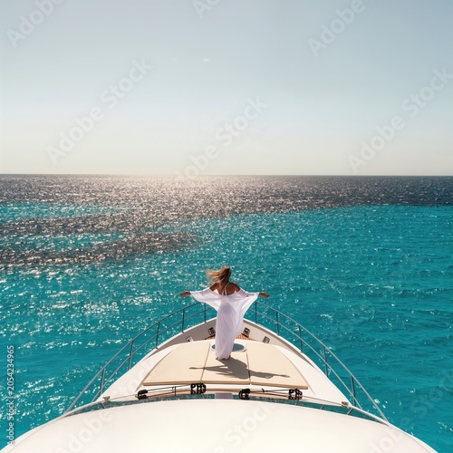 Young woman in white dress standing on yacht bow over turquoise sea