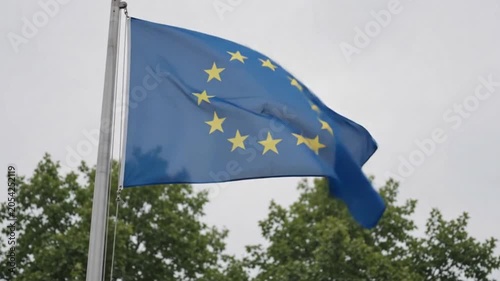 Flag of the european union waving in the wind against a cloudy sky and green trees in the background
