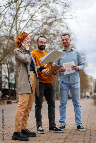 Group of three people discussing documents in a city park during a cloudy day with trees lining the background and a path in front of them
