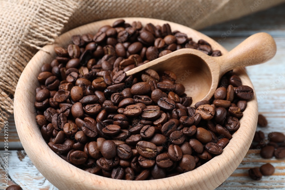Fototapeta premium Aromatic coffee beans in bowl, scoop and burlap fabric on wooden table, closeup