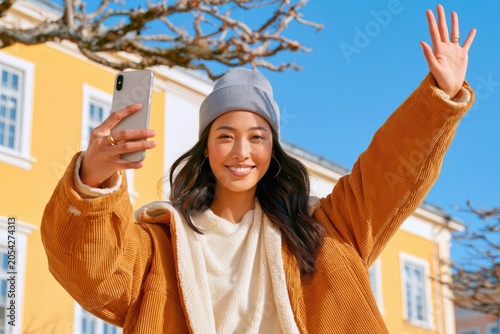 Young woman taking a smiling selfie outdoors with smartphone while waving in sunny urban setting