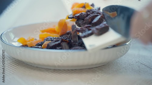 Hands using a knife to transfer chopped dried apricots and prunes from a cutting board into a plate.