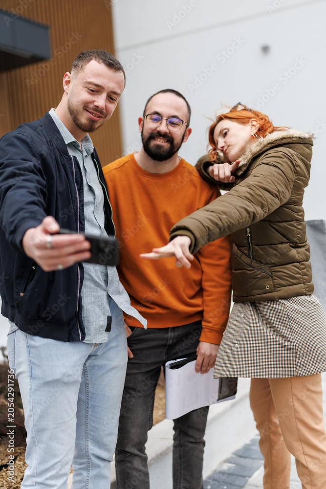 Obraz premium Three business professionals take a selfie outside an office building on a sunny day while dressed in casual attire and sharing smiles