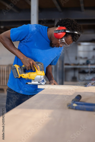 Carpenter is leaning on workshop bench using yellow jigsaw cutting wooden plank under bright lights