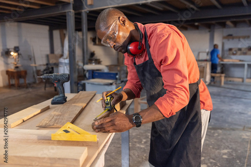 Foto Diverse coworkers wearing goggles marking wooden plank in shop with tape measure