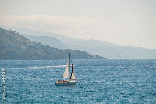 A lone sailboat cruising on the sparkling Mediterranean sea with a hazy, mountainous coastline in the background, French Riviera.