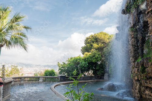 A scenic view of the waterfall at the Colline du Château (Castle Hill) park in Nice, France, featuring lush greenery, a stone pool, and a glimpse of the city in the background.