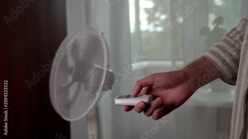 The hand of a young man in a linen shirt turns on an electric floor fan with a remote control during abnormal heat