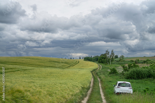 White car parked near a dirt road running through green wheat fields under a dramatic stormy sky.