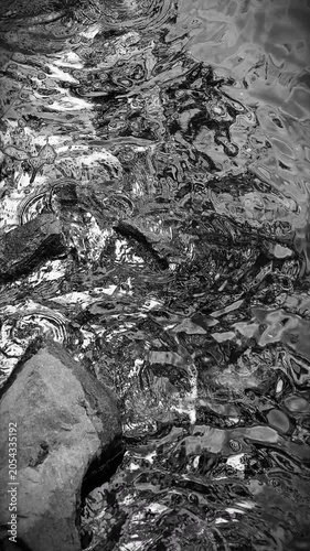 Black and white vertical format of shallow water rippling around big and small rocks, creating circles in a calm atmosphere. 