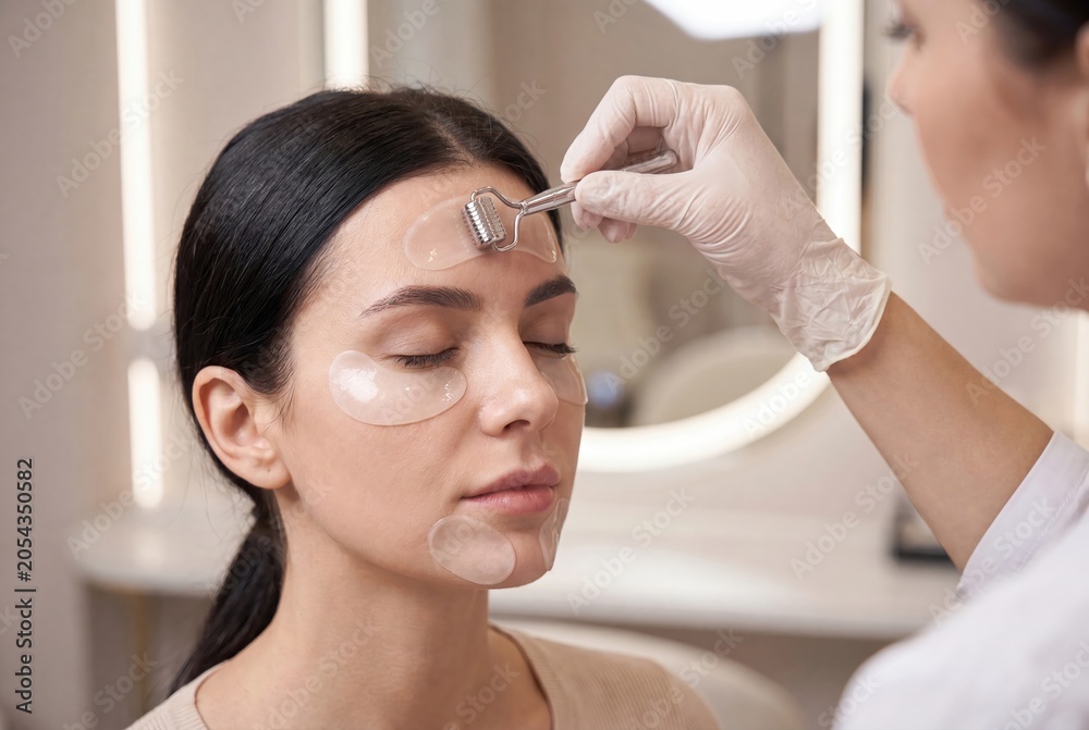 Naklejka premium Close-up of a woman receiving a facial treatment with a dermaroller and eye patches in a beauty salon.
