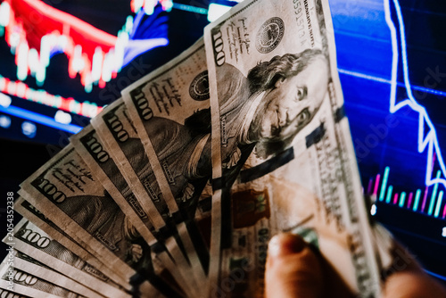 Hands holding US dollars currency in front of the stock exchange screen showing trading graphics.