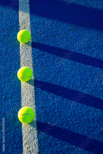 Close up view of tennis balls on a blue artificial grass surface with strong sunlight and dark shadows