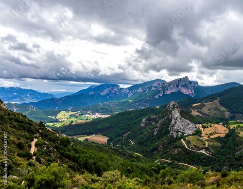 Vast panoramic vista with towering, jagged peaks and lush, rolling green hills under a dramatic, overcast sky