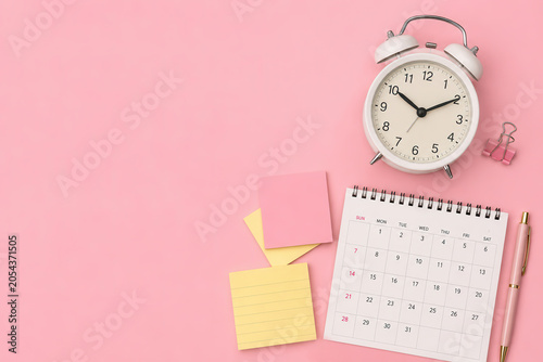 A clock shows the time beside a calendar and colorful sticky notes. A pen lies nearby on a pink background. This setup helps with planning and organizing tasks effectively.