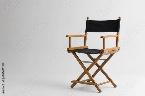 A wooden director's chair stands empty against a white backdrop. The chair has a black fabric seat and back. It is commonly used in film and photography settings.